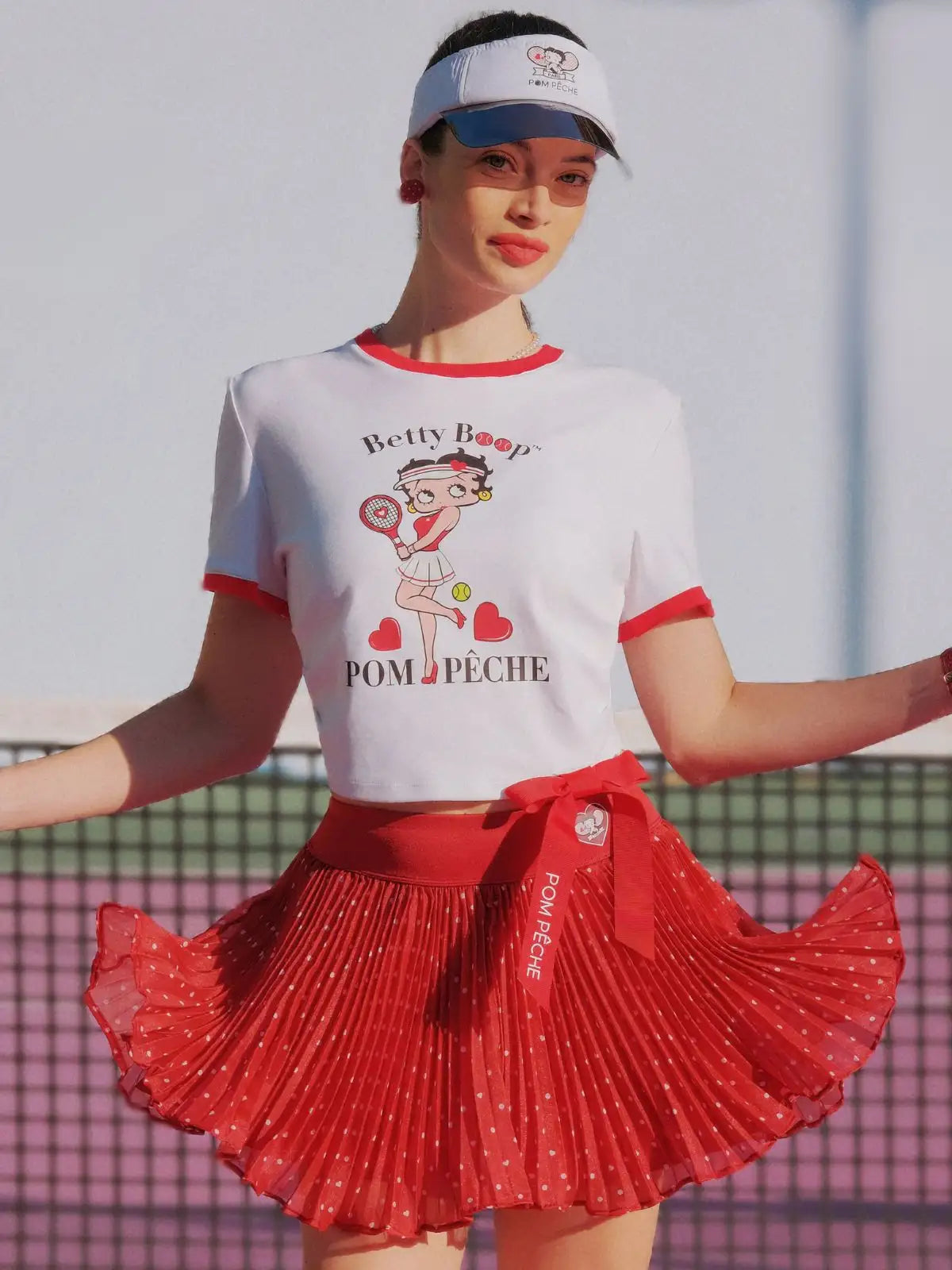 Woman wearing a white t-shirt with a graphic design and a red pleated skirt on a tennis court.