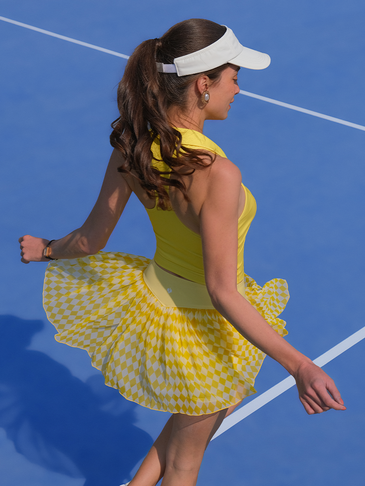 A woman wearing a yellow and white checkered print tennis skirt with a matching top and visor, playing tennis on a blue court.