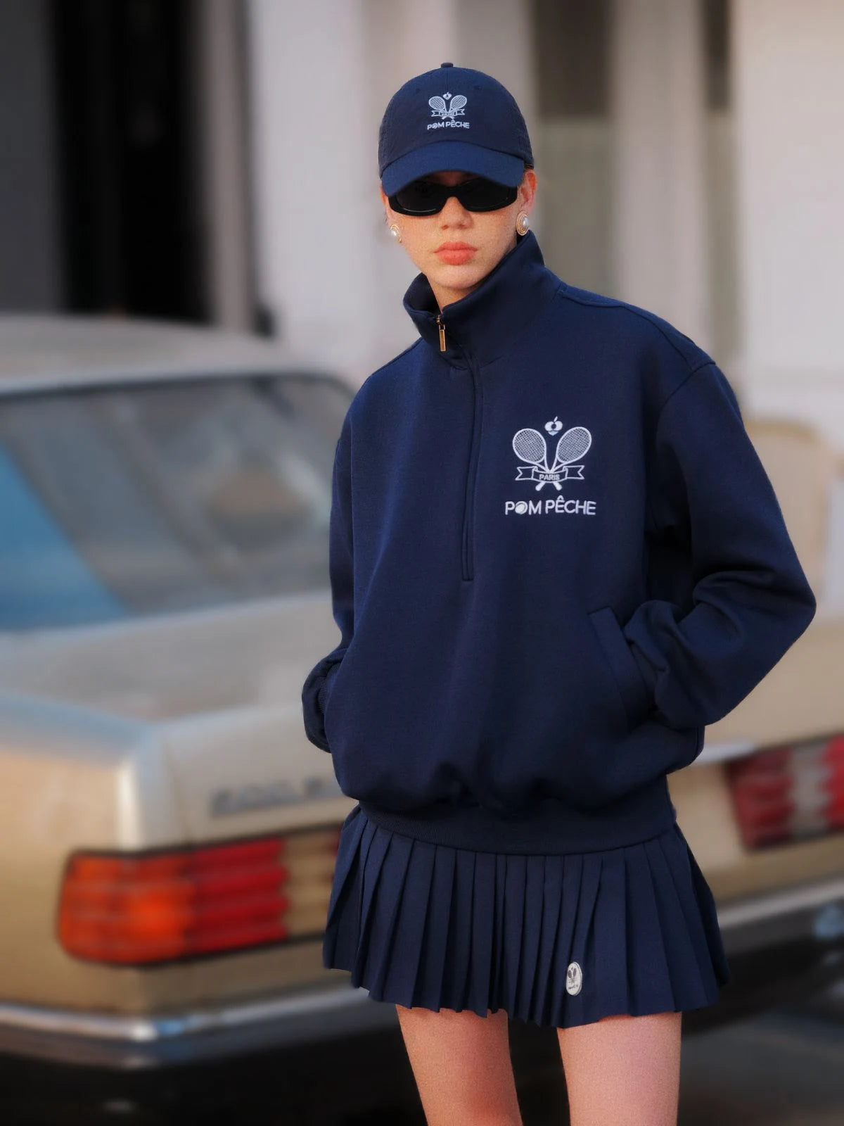 Person wearing a navy blue hoodie and skirt with a logo, standing in front of a vintage car.