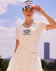 Pom Pêche x Betty Boop Tennis Tee in white – V-neck polo collar with Betty Boop Paris twin-racket graphic and Pom Pêche script, slim-fit short sleeve silhouette, styled with white Betty Boop Cake Skirt, Pom Pêche La Rose mirror visor and pearl necklace against Paris blue sky