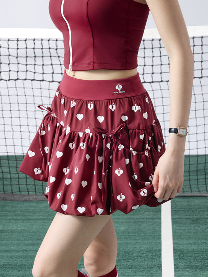 Person wearing a red athletic outfit with a skirt featuring white heart patterns on a tennis court.