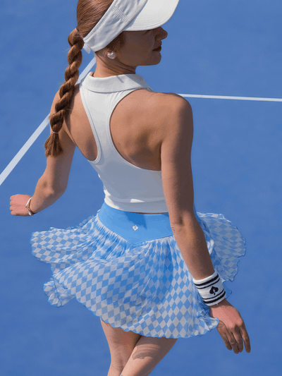 Woman wearing a white racerback top and the Argyle Mesh Tennis Skirt in Blue spinning on a blue tennis court, showcasing the lightweight argyle mesh fabric.