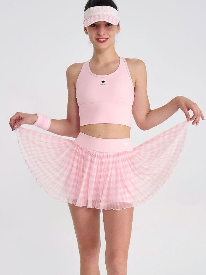 woman wearing a light pink argyle mesh pleated tennis skirt and matching sports bra, holding the skirt out to show the flowy layers in a studio setting