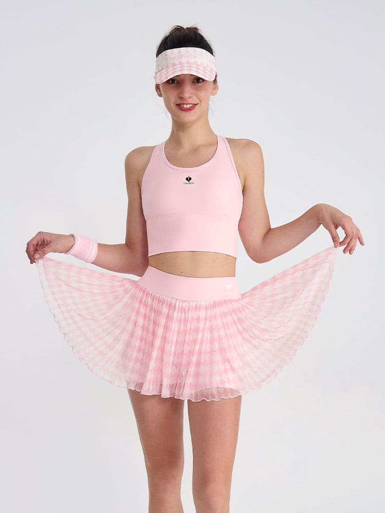 woman wearing a light pink argyle mesh pleated tennis skirt and matching sports bra, holding the skirt out to show the flowy layers in a studio setting