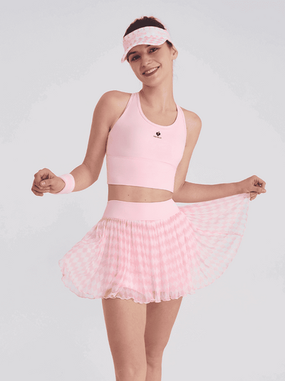 female tennis player in a light pink argyle mesh tennis skirt posing with the pleated hem spread out, styled with a pink athletic top and visor