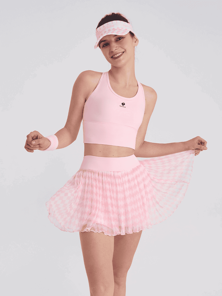 female tennis player in a light pink argyle mesh tennis skirt posing with the pleated hem spread out, styled with a pink athletic top and visor