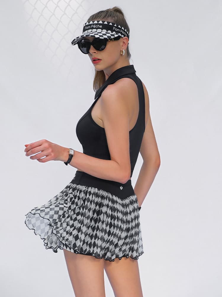 A woman walks in front of a white backdrop wearing a black and white argyle mesh tennis skirt and black athletic top.