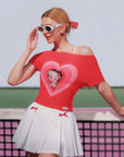 Woman wearing a red off-shoulder top with a heart design and white skirt on a tennis court.
