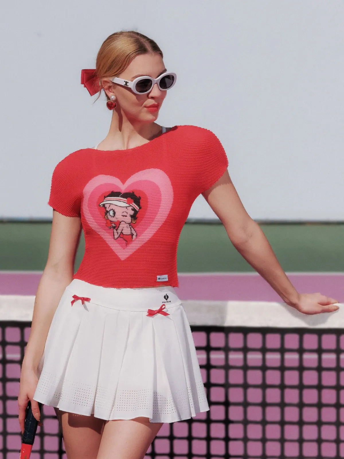 Woman wearing a red top with a heart design and white skirt on a tennis court.