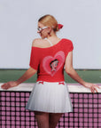 Woman wearing a red top with a heart design and white skirt, standing on a tennis court.