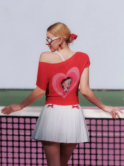 Woman wearing a red top with a heart design and white skirt, standing on a tennis court.