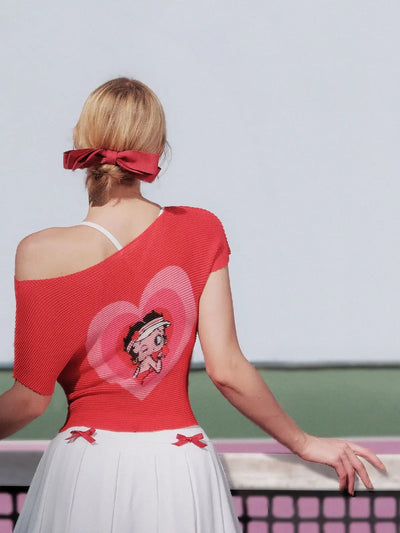 Woman wearing a red top with a heart design and white skirt, standing against a light gray wall.