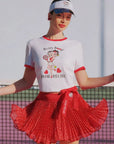 Woman wearing a white t-shirt with a graphic design and a red pleated skirt on a tennis court.