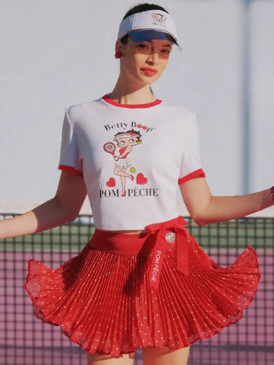 Woman wearing a white t-shirt with a graphic design and a red pleated skirt on a tennis court.