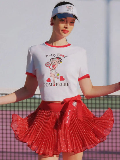 Woman wearing a white t-shirt with a graphic design and a red pleated skirt on a tennis court.