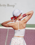 Woman on a tennis court wearing a white top and skirt with red accents, with 'Betty Boop' branding in the background.
