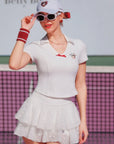 Woman in a white tennis outfit with a skirt and top, standing on a tennis court.