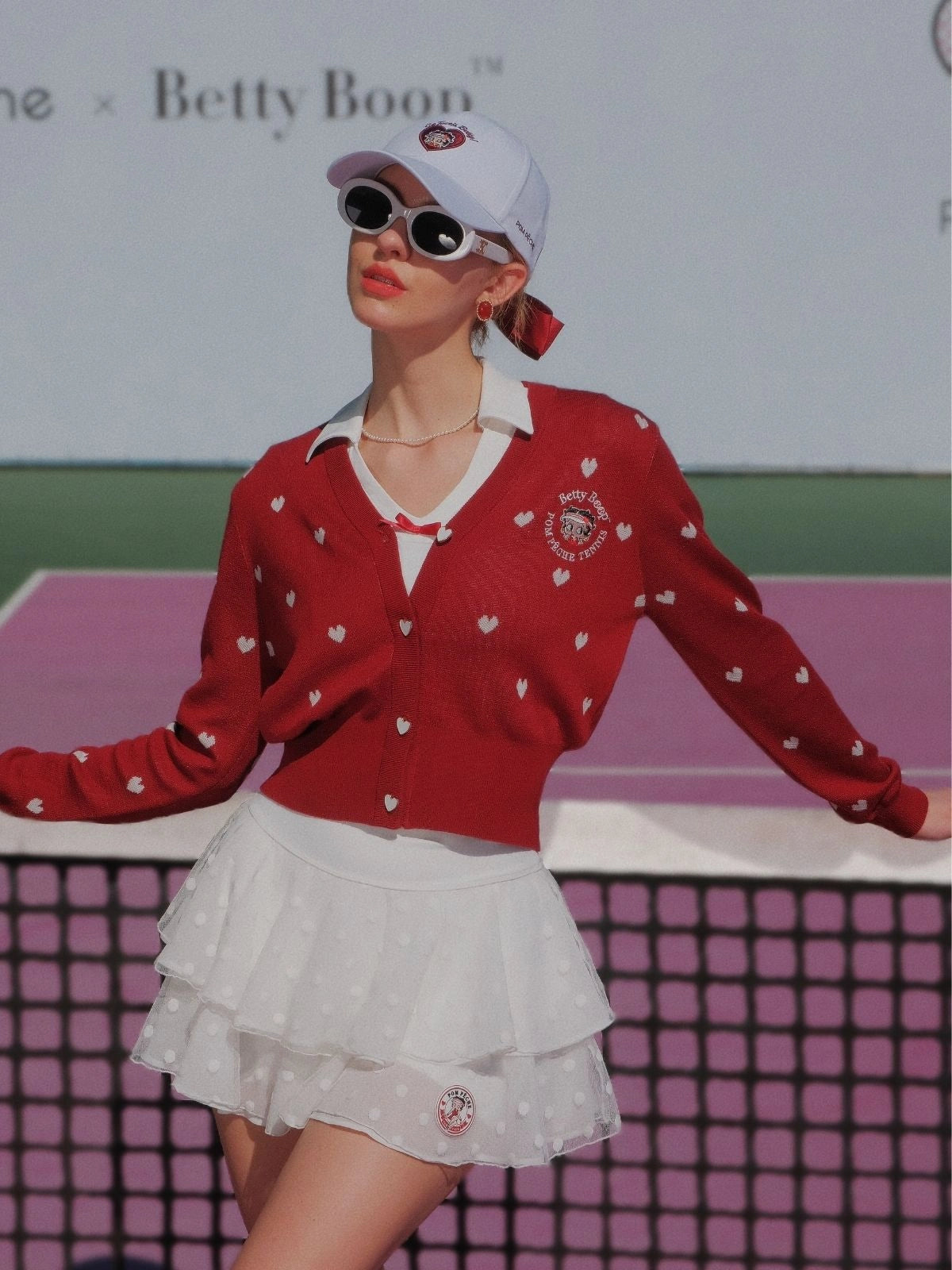 Person wearing a red sweater with white heart patterns and a white skirt on a tennis court.
