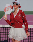 Woman in red cardigan and white skirt holding a paddle on a tennis court