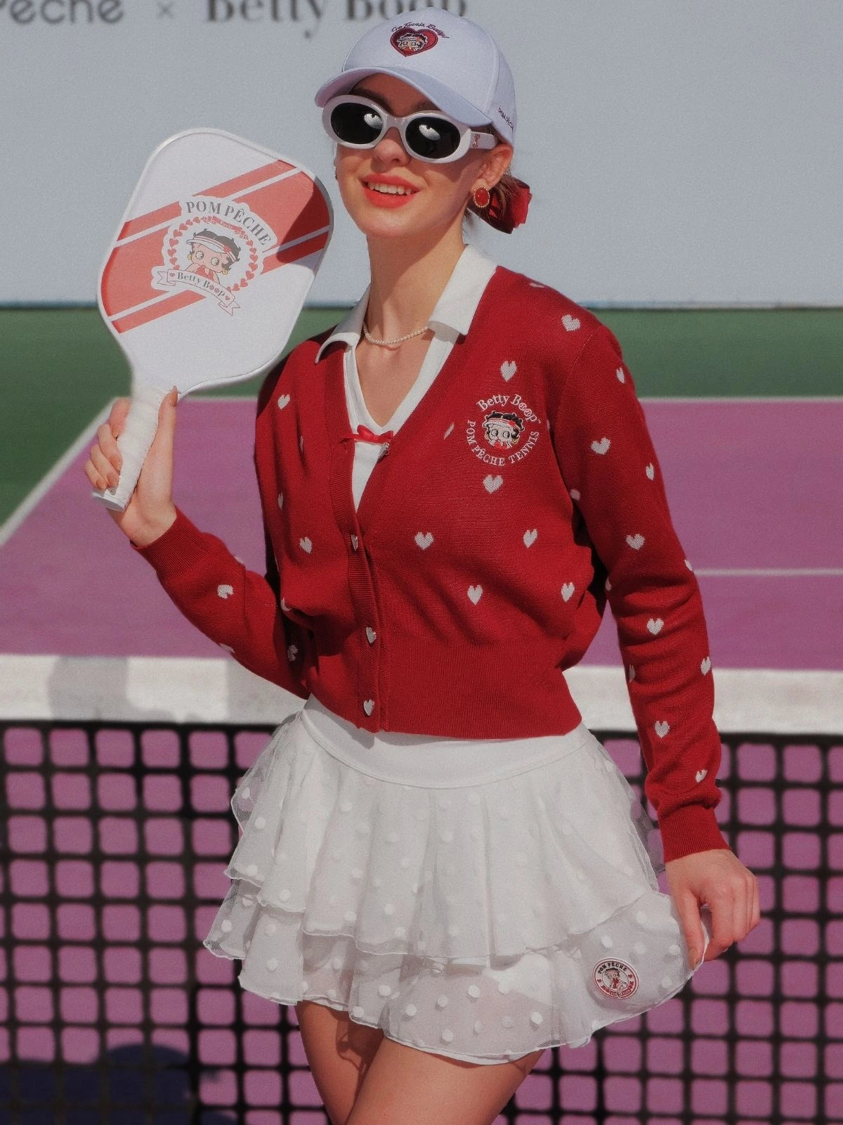 Woman in red cardigan and white skirt holding a paddle on a tennis court