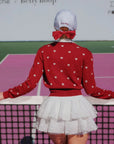 Person wearing a red sweater with white heart patterns and a white skirt on a tennis court.