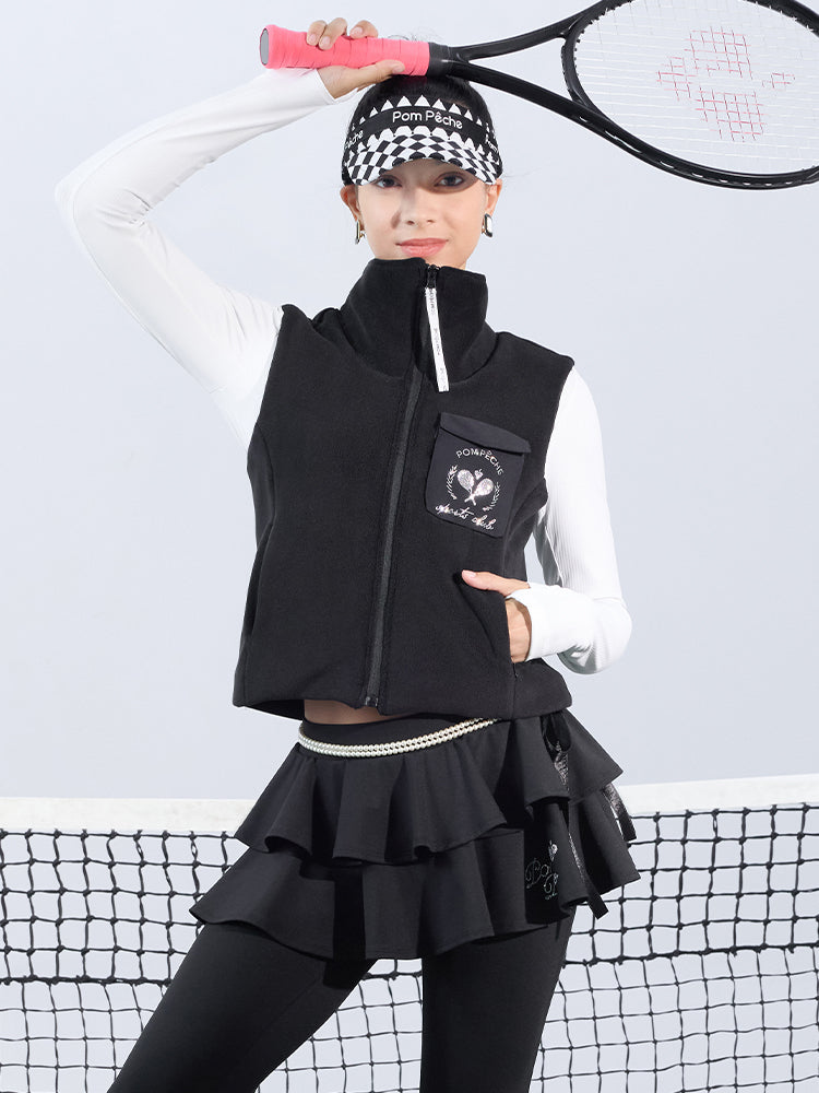 Warm black fleece vest with high-neck zip design and embroidered chest pocket, styled on the tennis court.