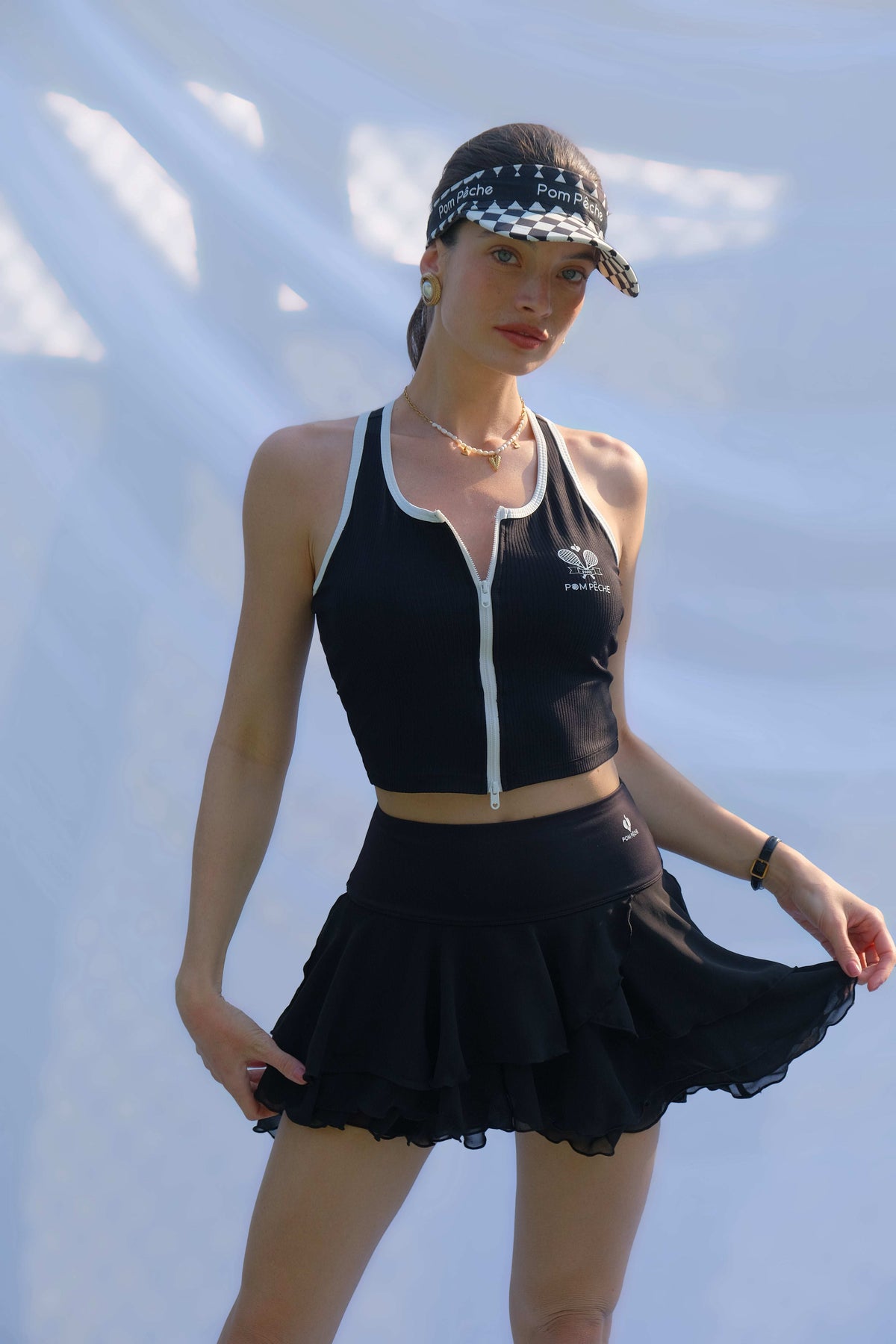 A woman poses on the tennis court holding the hem of her black ruffle tennis skirt while wearing a black zip-front athletic top.