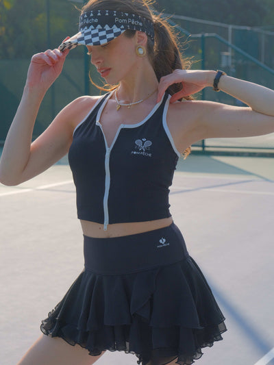 A woman adjusts her checkered visor while wearing a black ruffle tennis skirt and matching sports top on an outdoor tennis court.