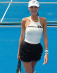 Woman on a tennis court wearing a white top and black skirt with a visor.