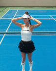Woman on a tennis court wearing a white top, black skirt, and white visor.
