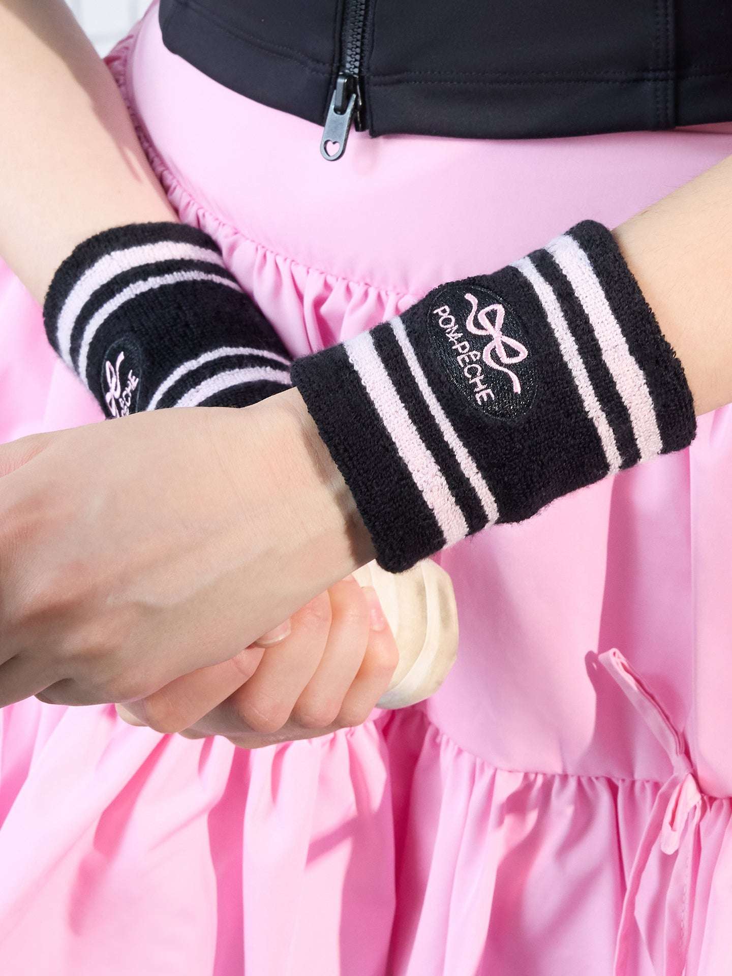 Black tennis wristbands with soft pink stripes, featuring embroidered bow and logo details.