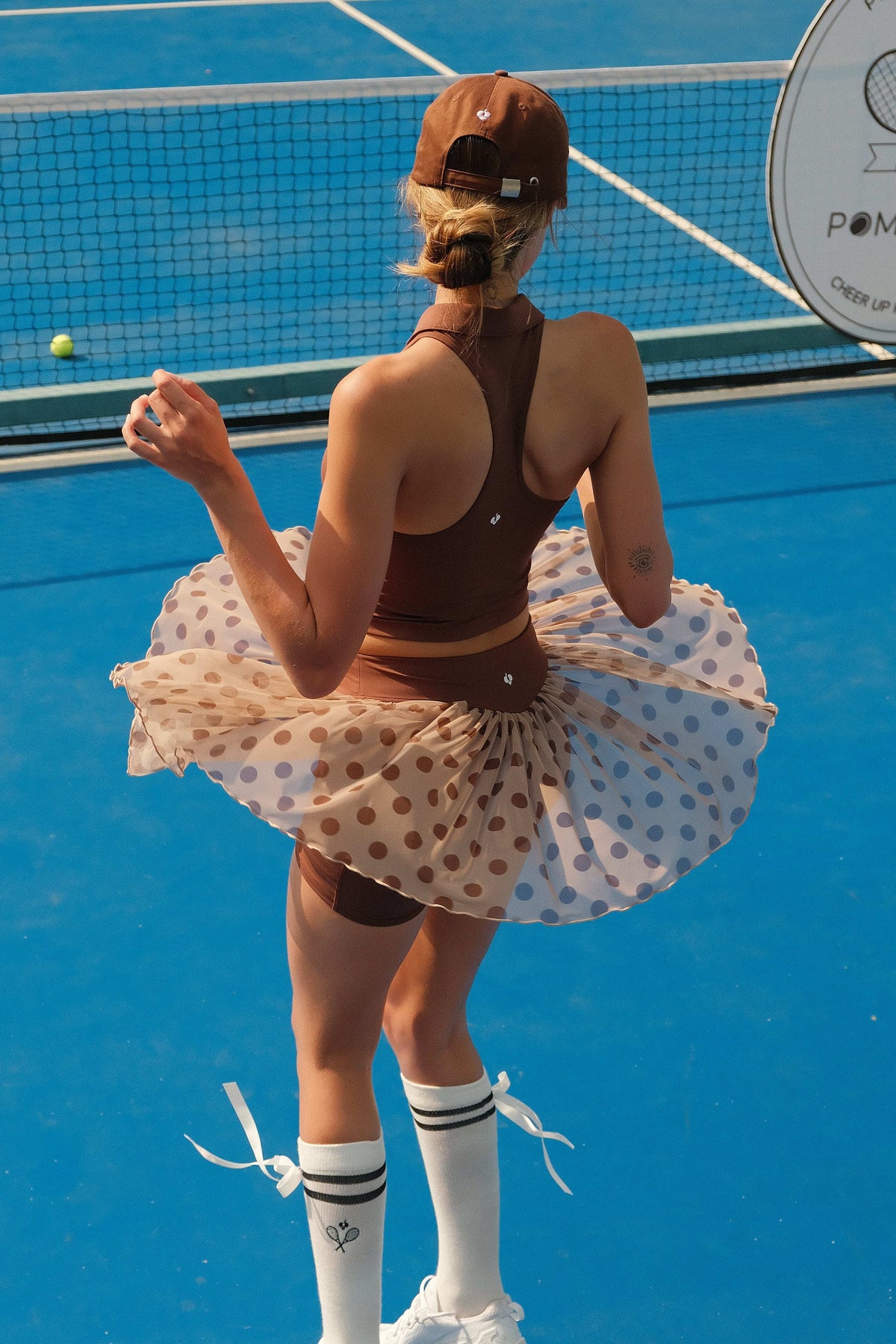 A woman in a brown polka dot mesh tennis skirt spinning on a blue tennis court, showcasing the lightweight athletic mesh fabric and built-in shorts.