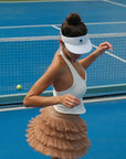Woman on a tennis court wearing a white visor and beige skirt, preparing to serve.