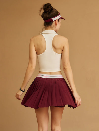 Woman wearing a white crop top and wine red tennis skirt with a visor on a beige background