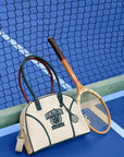 Cream dome tennis bag with contrast handles and Pom Pêche logo, styled on a tennis court.