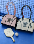 Cream dome tennis bag with contrast handles and Pom Pêche logo, styled on a tennis court.