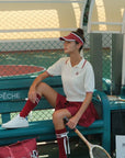 Woman sitting on a bench with a tennis racket, wearing a red and white outfit, in an outdoor setting.