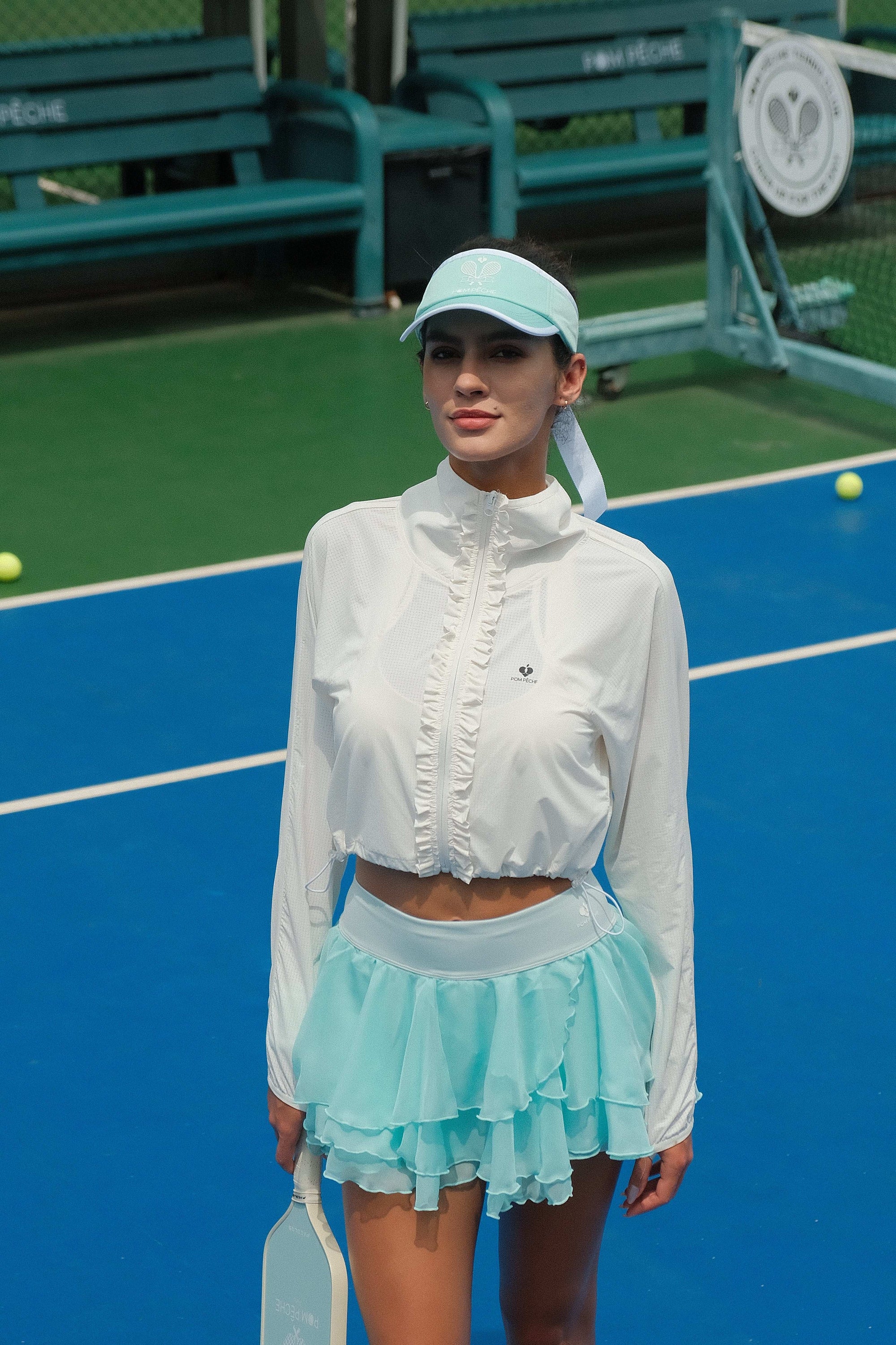 White cropped jacket with ruffled zip-front detail and long sleeves, styled on the tennis court.