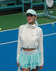 White cropped jacket with ruffled zip-front detail and long sleeves, styled on the tennis court.