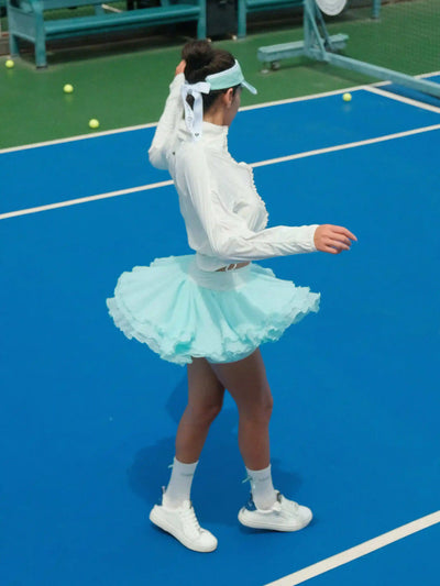 Person on a tennis court wearing a white outfit with a light blue tutu skirt.
