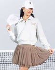 Grey sweatshirt with quarter-zip design and embroidered logo, styled on the tennis court for a chic, sporty look.