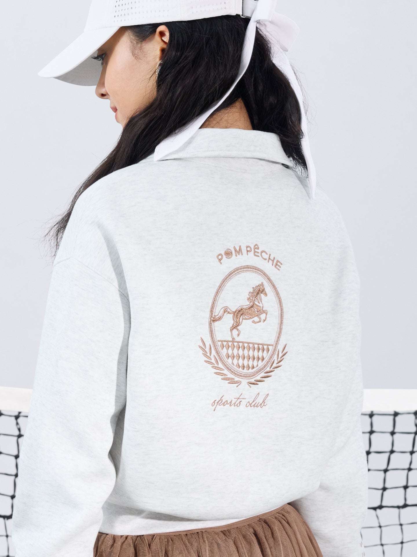 Gray sweatshirt with quarter-zip design and embroidered logo, paired with a tulle skirt, styled on the tennis court for a chic, sporty look.
