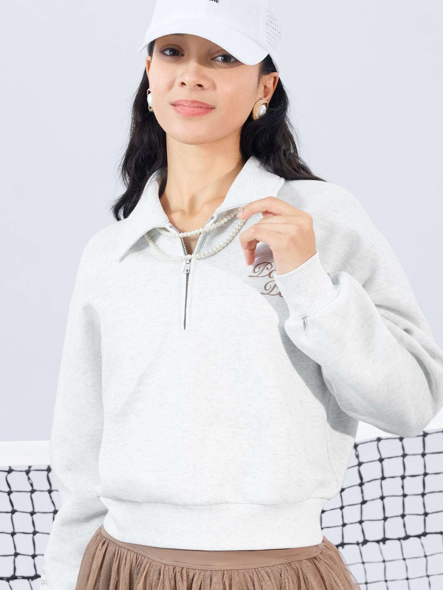 Gray sweatshirt with quarter-zip design and embroidered logo, paired with a tulle skirt, styled on the tennis court for a chic, sporty look.