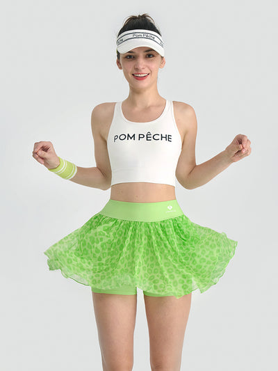 A woman wearing a green leopard print mesh tennis skirt poses on court, showing the lightweight athletic fabric and high-waisted fit.