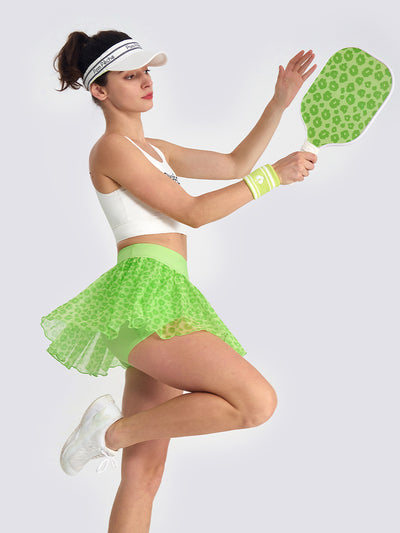 A tennis player jumps while wearing a green leopard print tennis skirt, highlighting the flowy mesh layers and built-in shorts for sports performance.