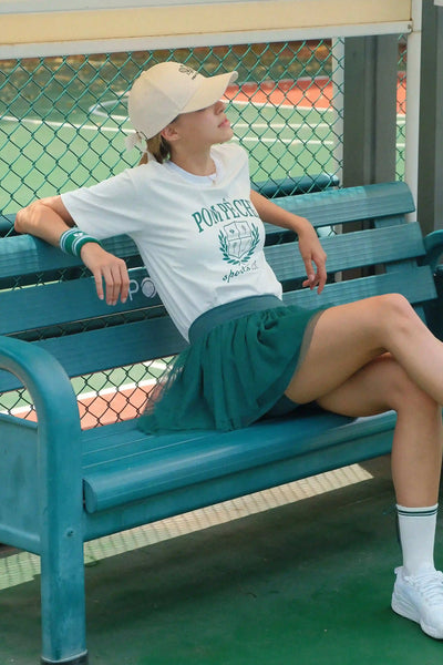 Person sitting on a blue bench wearing a white t-shirt with green text and a green skirt on a tennis court.
