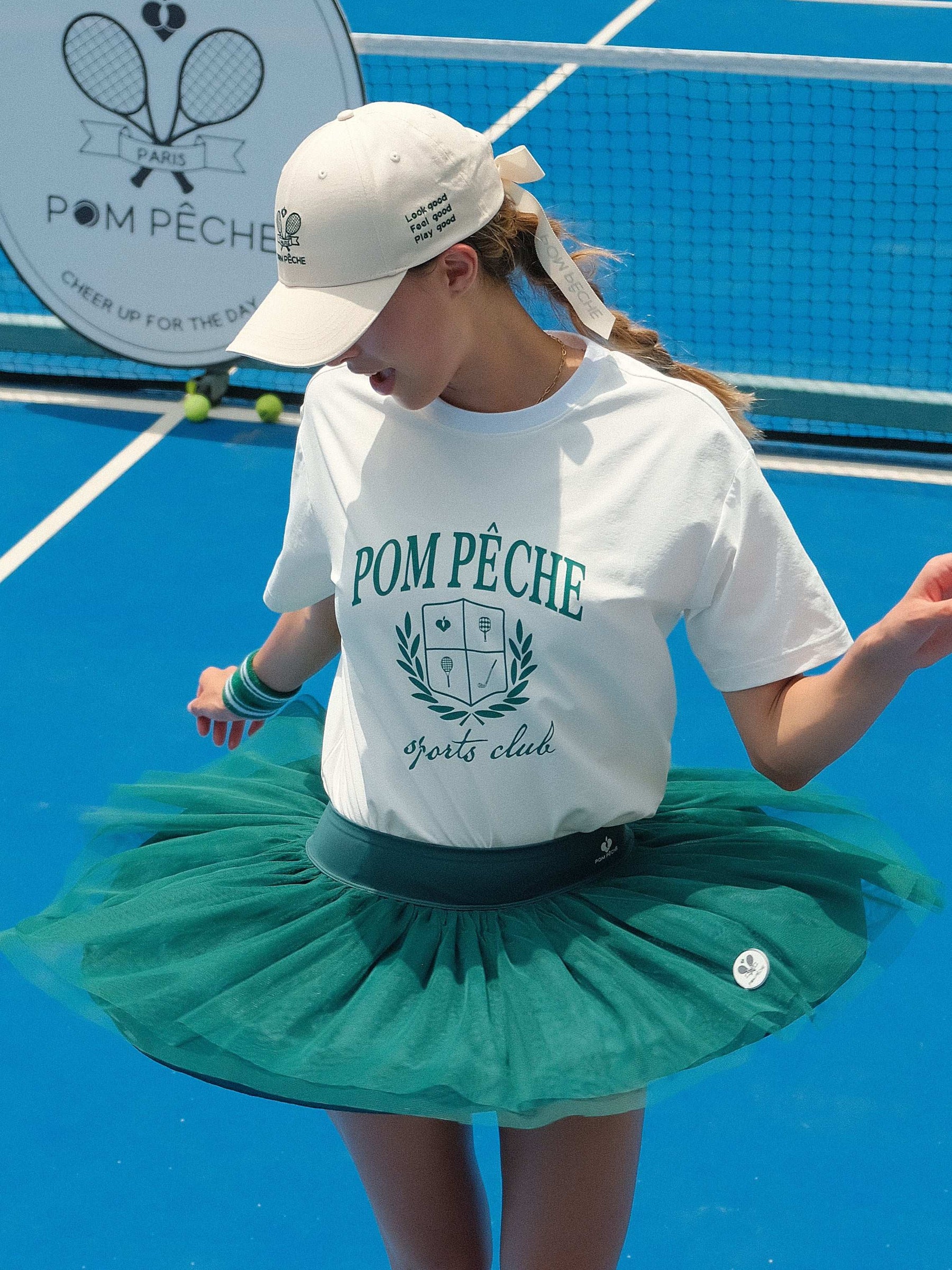 Model wearing a green tulle puff tennis skirt on blue court, showing airy layered volume and a high-waisted athletic tennis skirt look.