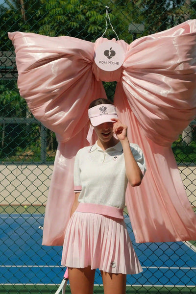 Person in tennis attire with a large pink bow and visible brand logo on a tennis court.