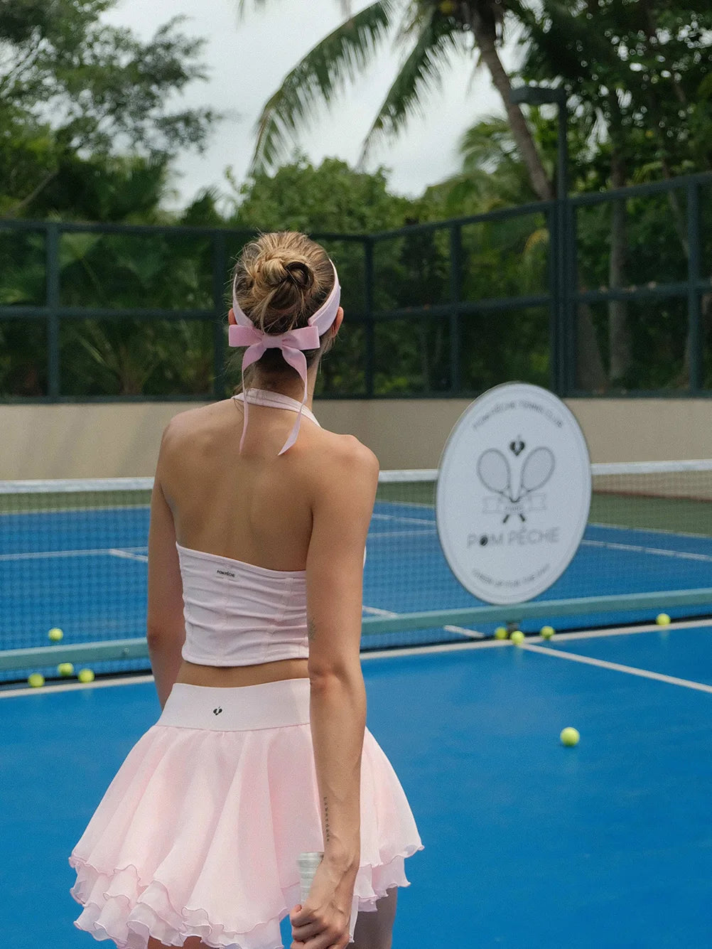Woman in a white top and pink skirt on a tennis court with palm trees in the background