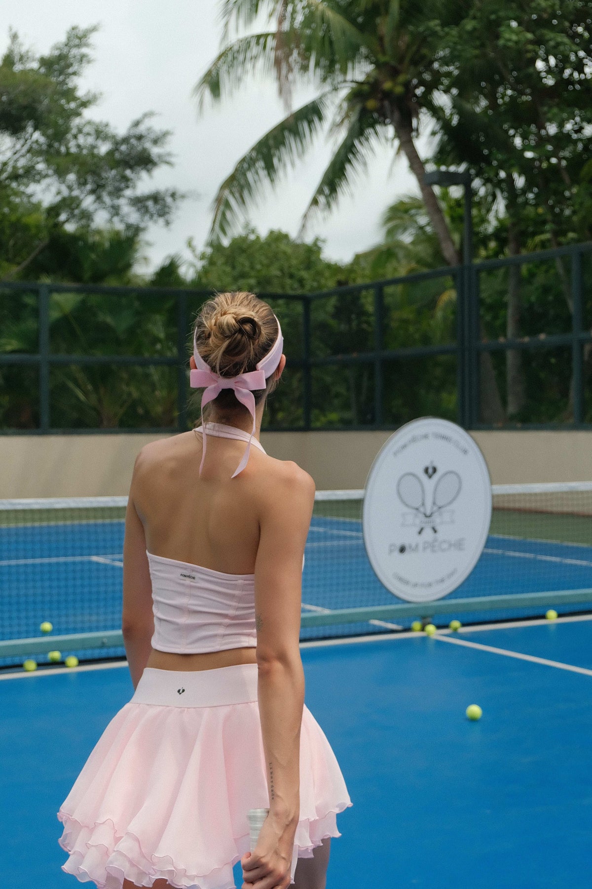A woman walks toward the net on a blue tennis court wearing a pink layered tennis skirt and a ribbon visor.